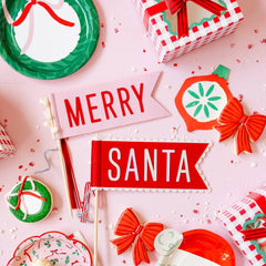 Christmas-themed party supplies including a 'Merry Santa' sign, cookies, and decorative elements on a pink background.